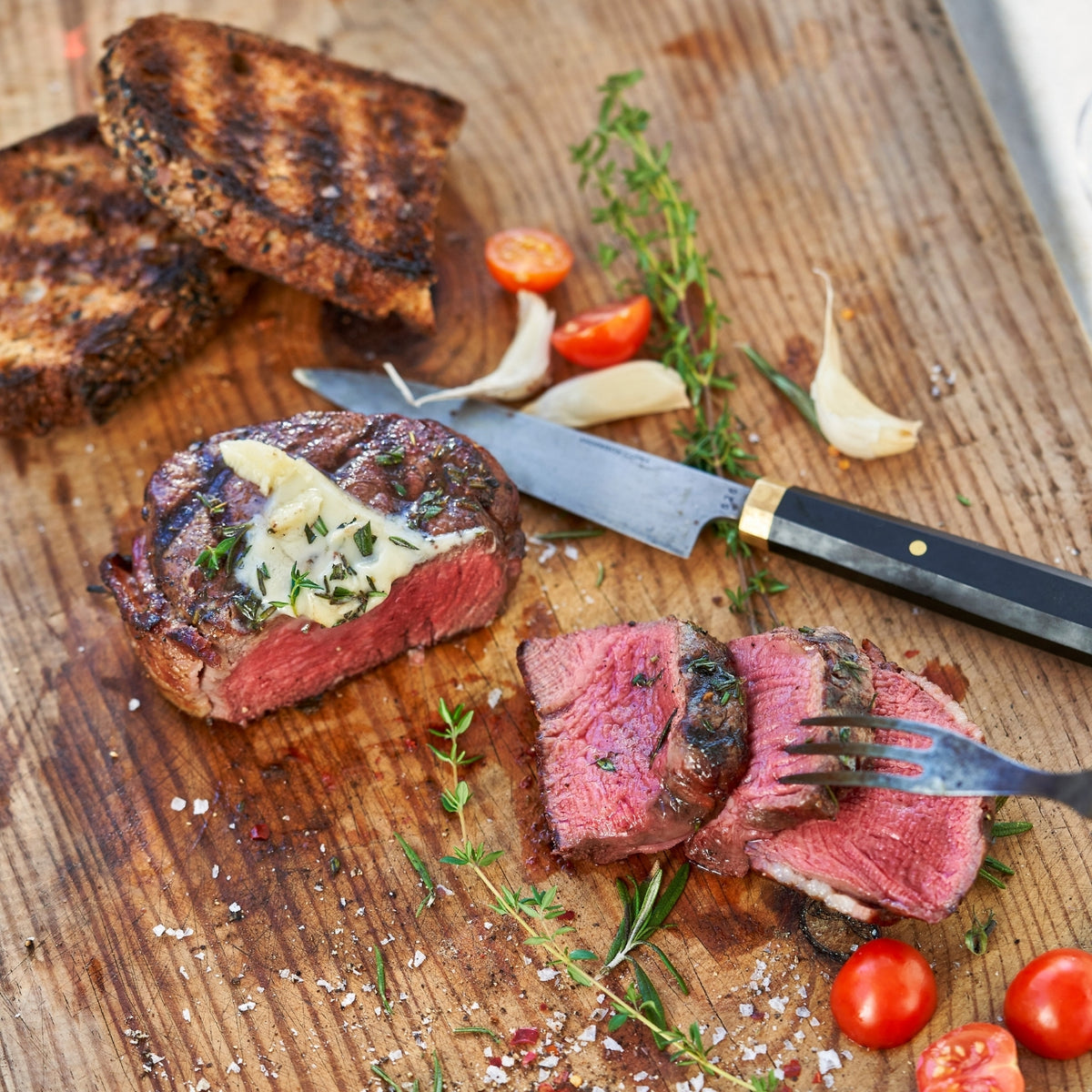 Sliced steak with garlic and herbs on a wooden cutting board with a knife and fork.