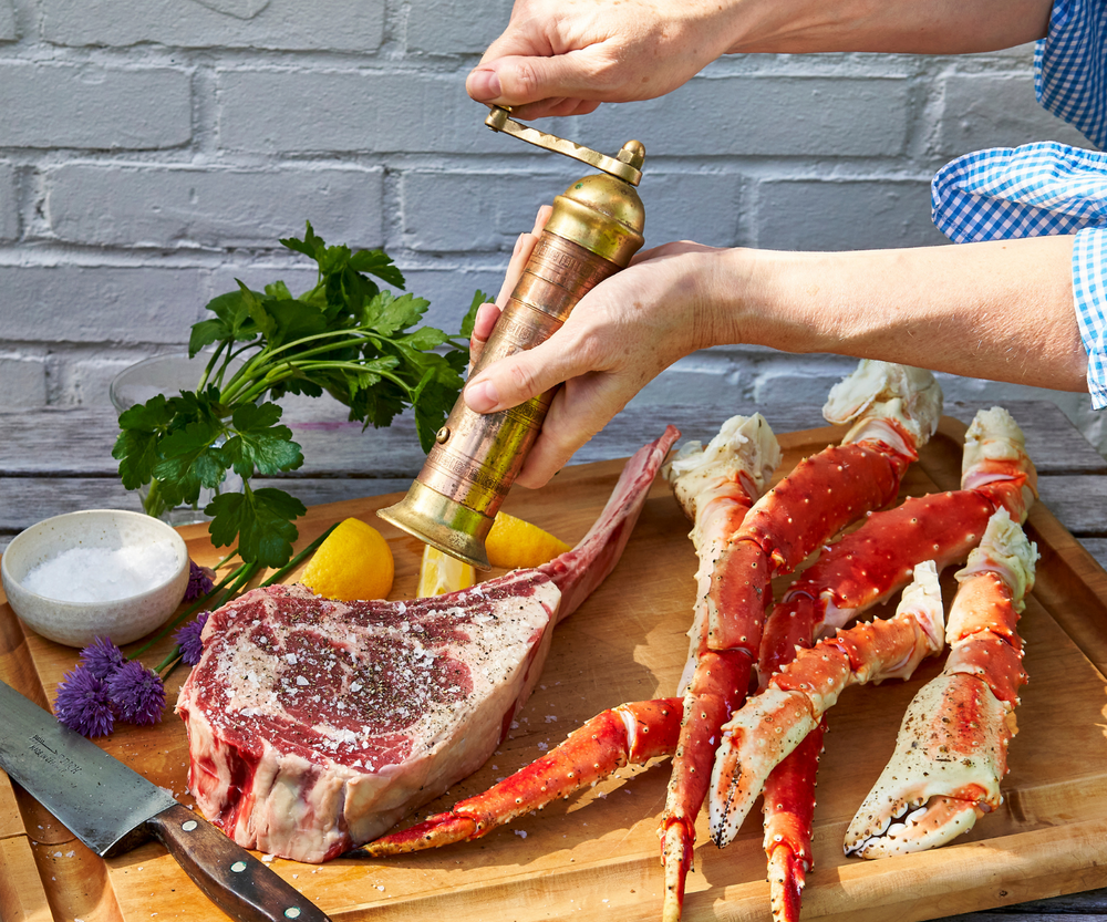 Person seasoning a steak with a pepper grinder on a wooden cutting board with crab legs.