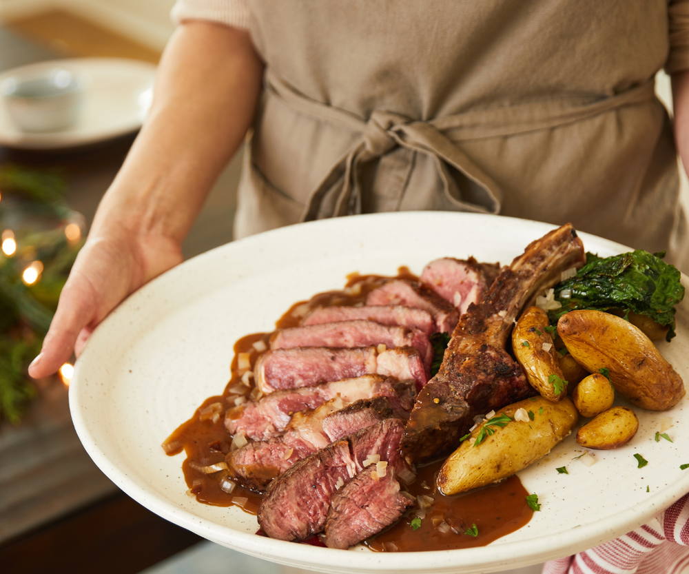 Person holding a plate of sliced steak with potatoes and greens in a home setting.