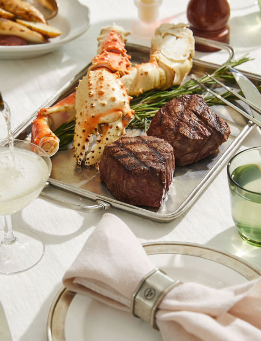 Tray of cooked crab legs and steaks on a dining table with glasses and cutlery.
