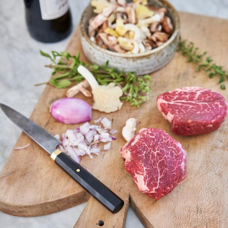 Raw steaks on a wooden cutting board with a knife, chopped onions, and a bowl of mushrooms.