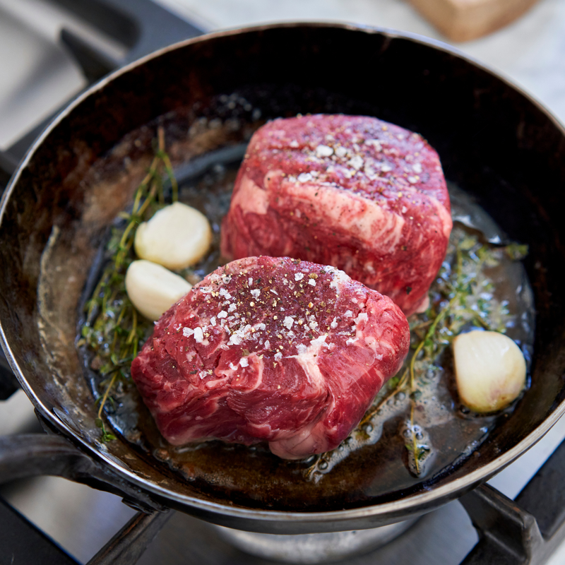 Two raw steaks with garlic and herbs in a cast iron skillet on a stovetop.