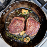 Two seared steaks in a frying pan with herbs and garlic on a stove.