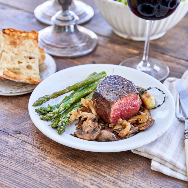 Steak with asparagus and mushrooms on a plate with bread and wine in the background.