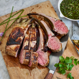Sliced roasted lamb on a wooden cutting board with herbs and a bowl of green sauce.