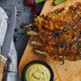Roasted rack of lamb with herbs on a wooden cutting board, accompanied by a small bowl of mustard sauce.