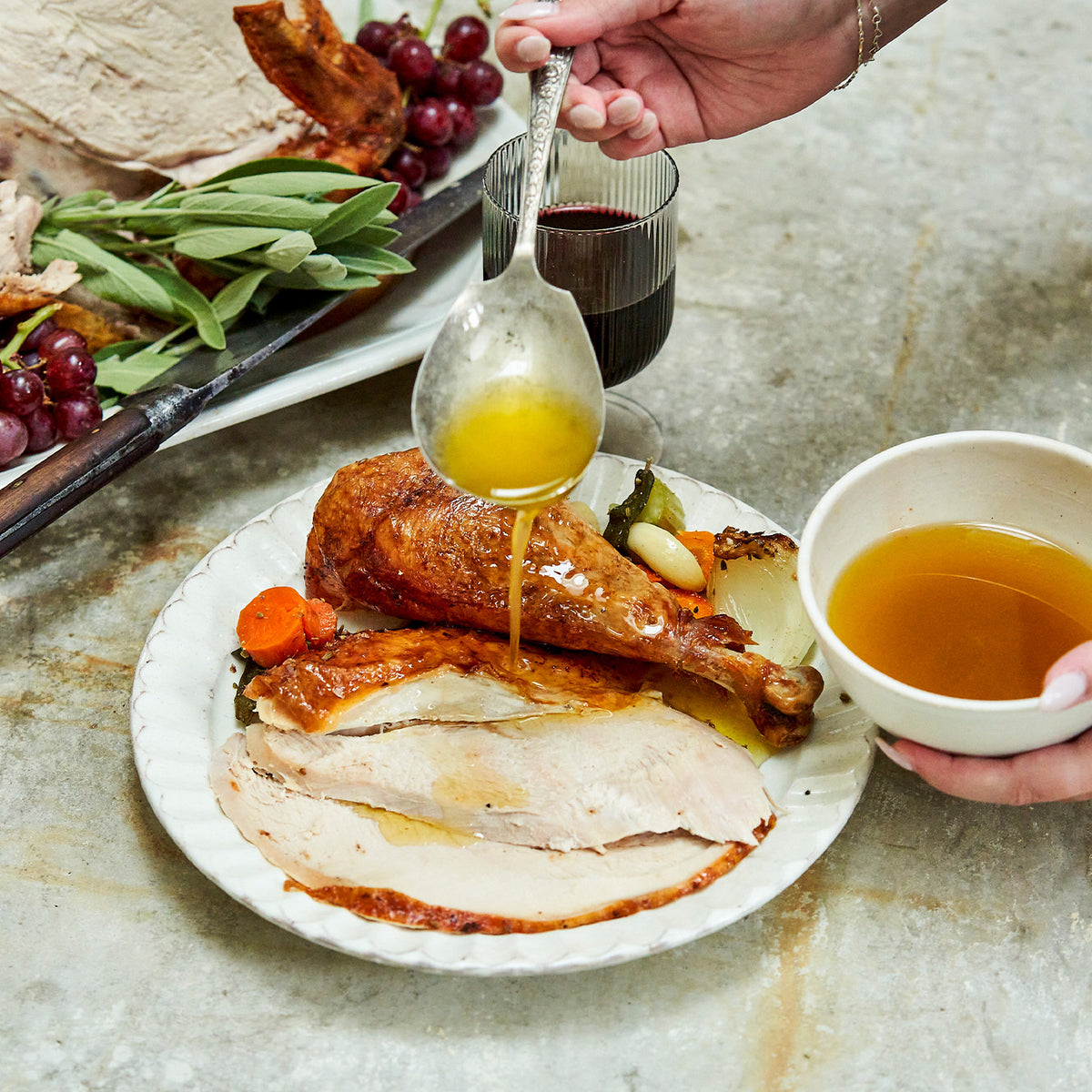 Person pouring sauce over roasted turkey on a plate with a bowl of sauce and wine glass in the background.