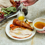 Person pouring sauce over roasted turkey on a plate with a bowl of sauce and wine glass in the background.