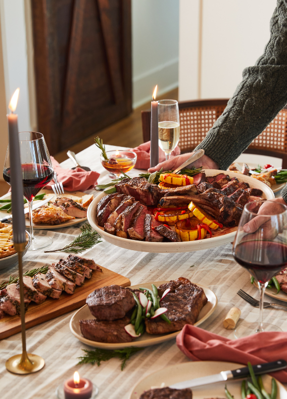 Dinner table set with grilled meats, vegetables, and wine glasses in a cozy indoor setting.