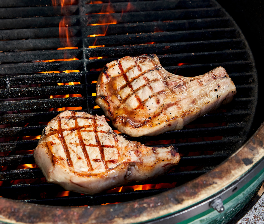 Two grilled pork chops on a barbecue grill with flames below.