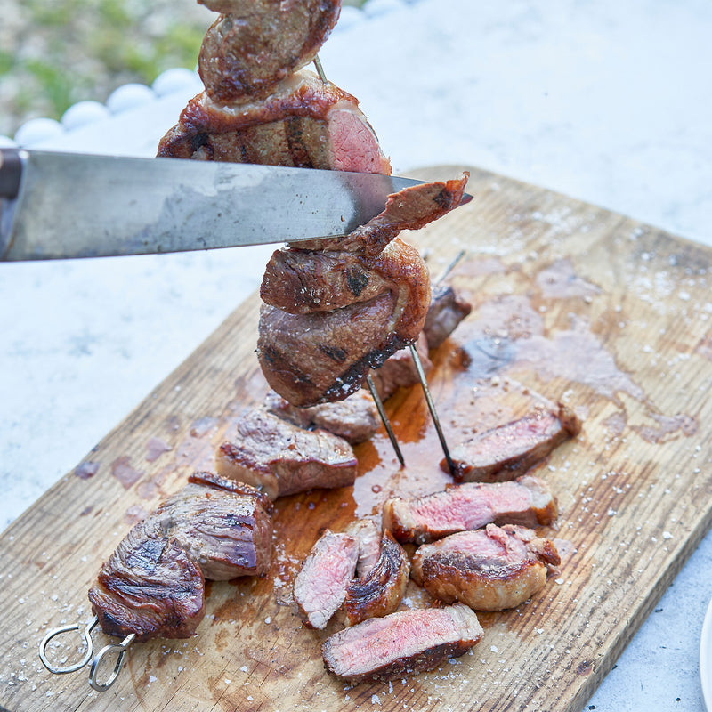Grilled meat skewers on a wooden cutting board with a knife in the background.