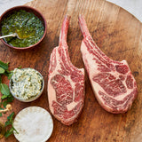 Two raw ribeye steaks on a wooden cutting board with bowls of green herb sauce and garlic compound butter.