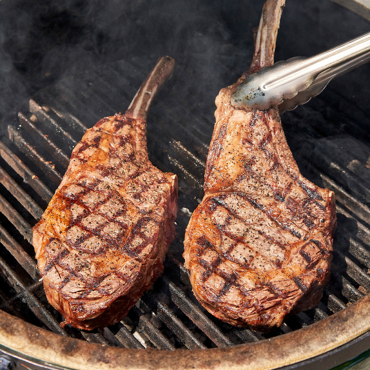 Two grilled steak pieces on a barbecue grill with tongs.