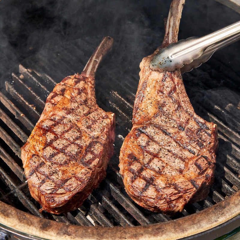 Two grilled steak pieces on a barbecue grill with tongs.
