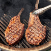 Two grilled steak pieces on a barbecue grill with tongs.