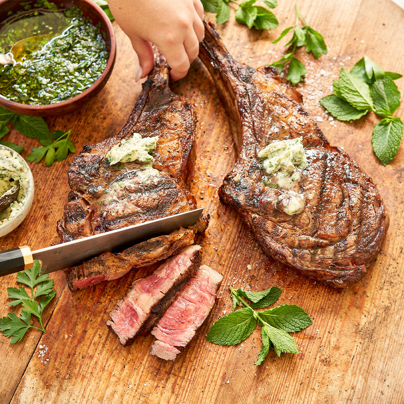 Tomahawk Ribeye being cut on a wooden cutting board with herbs and a bowl of green sauce.