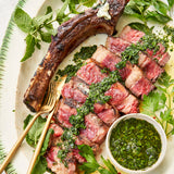 Grilled steak with chimichurri sauce on a plate with herbs and a gold fork.