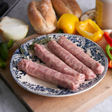 Three raw sausages on a decorative plate with vegetables and bread in the background.