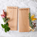 Two wooden cedar planks with 'Market House' branding, surrounded by herbs and a lemon on a textured white background.