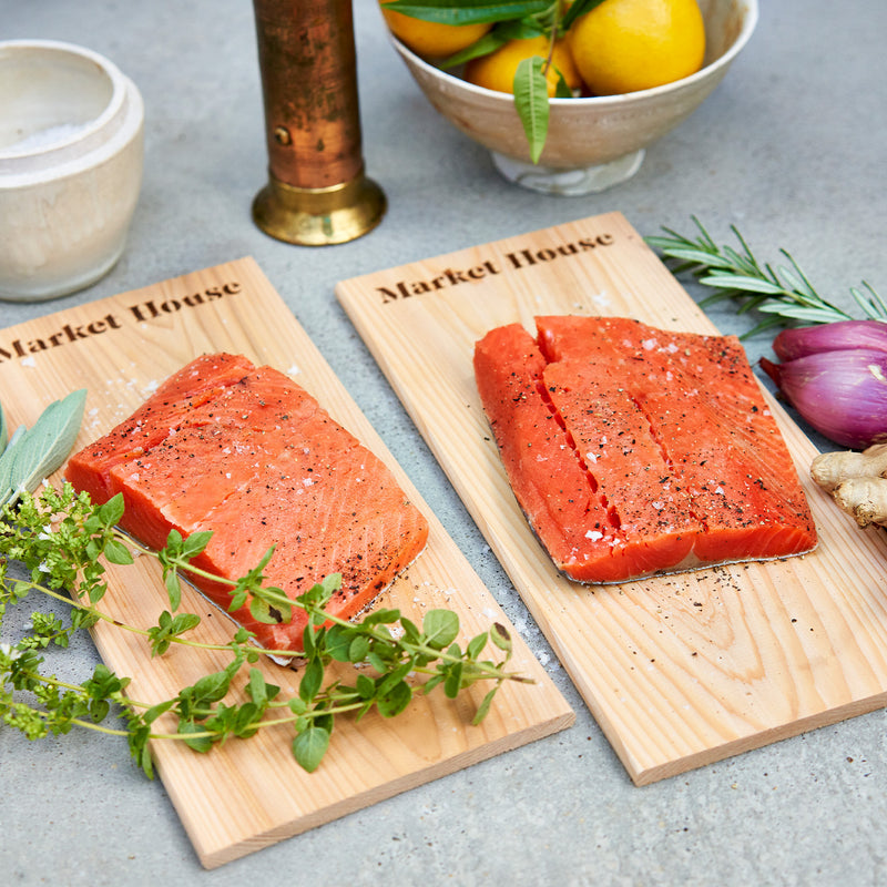 Two pieces of salmon on wooden cedar planks with herbs and a bowl of lemons in the background.