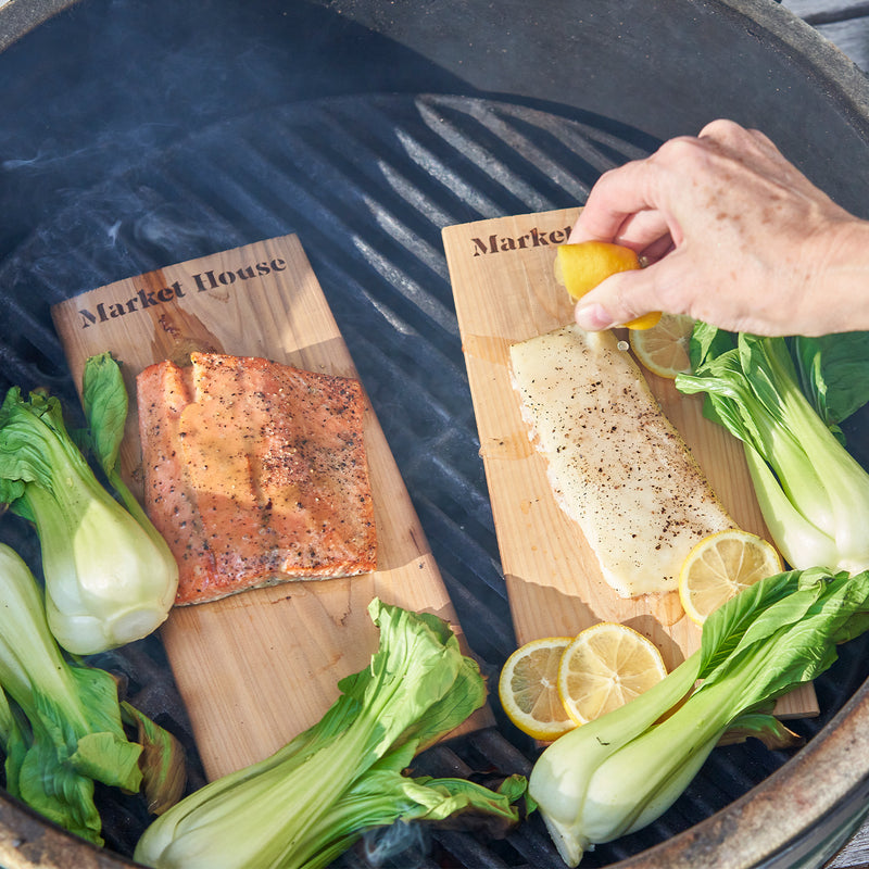 Person seasoning fish on a wooden board with lemons and bok choy on a grill.