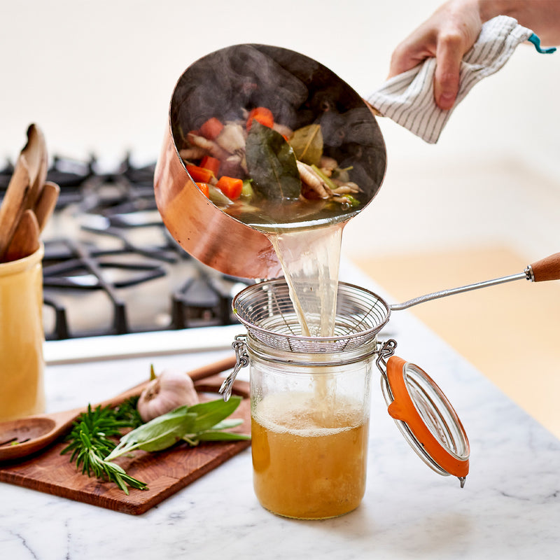 Person pouring liquid from a copper pot into a jar through a strainer on a kitchen counter.