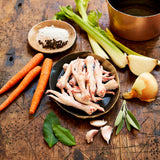 Raw chicken feet on a wooden surface with carrots, celery, onions, and herbs.