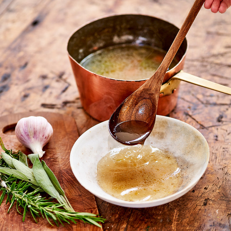 Wooden spoon transferring bone broth from a bowl to a copper pot on a wooden surface with herbs and garlic.