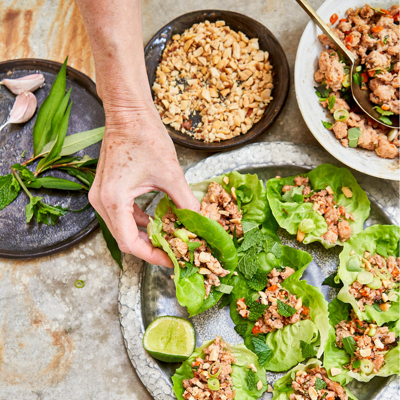 Hand holding a lettuce wrap with a plate of similar wraps and ingredients on a stone surface.