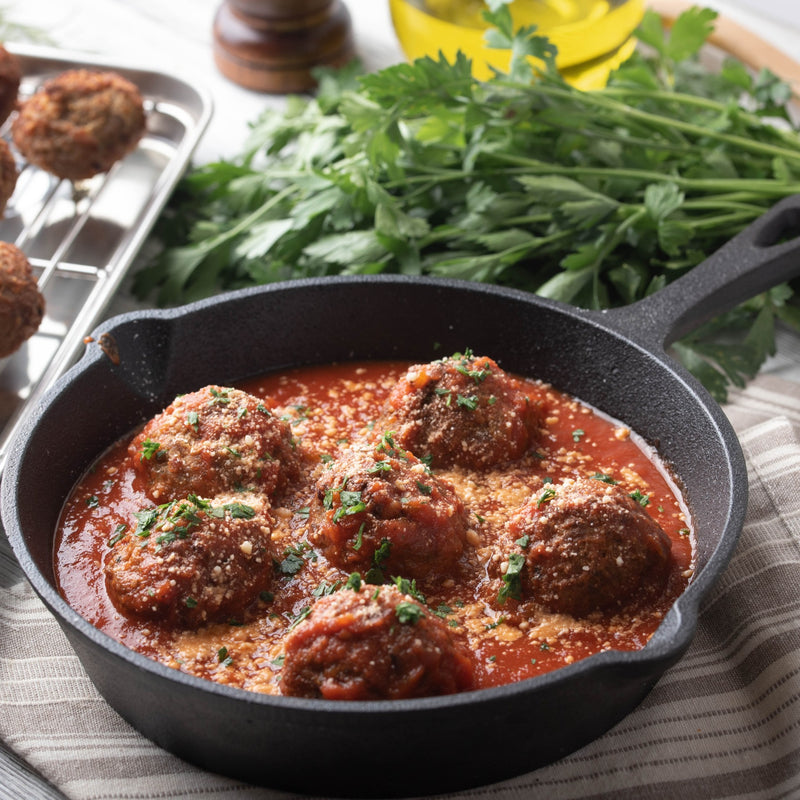 Skillet with meatballs in tomato sauce, fresh parsley, and oil in the background.