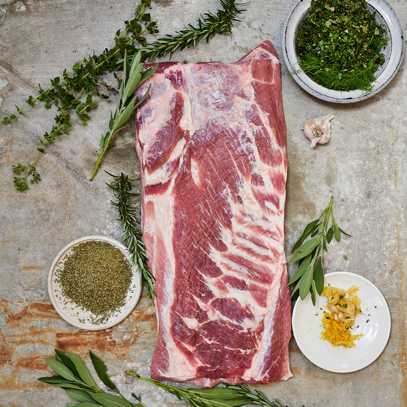 Raw Boneless, Skin-On Whole Pork Belly with herbs and spices on a stone surface