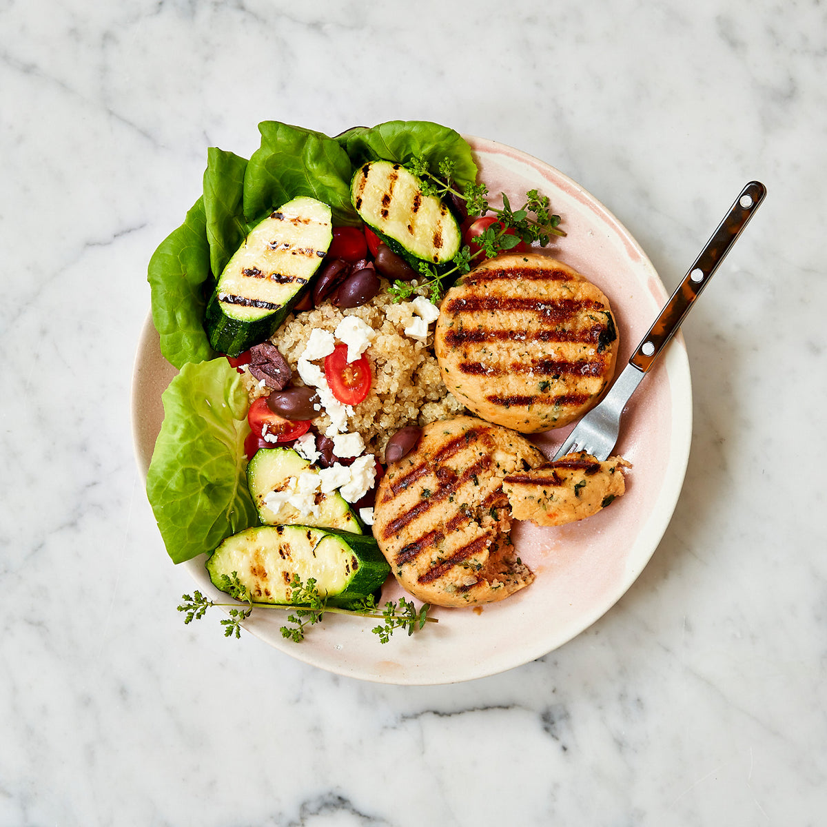 Plated dish with grilled salmon patties, quinoa, vegetables, and a fork on a marble background