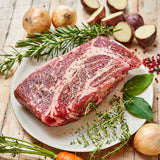 Raw chuck roast on a plate with herbs and vegetables on a wooden surface