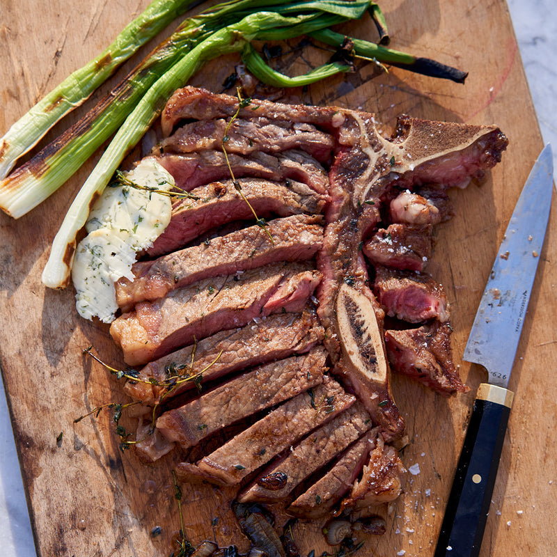 Sliced T-bone steak with green onions on a wooden cutting board with a knife.