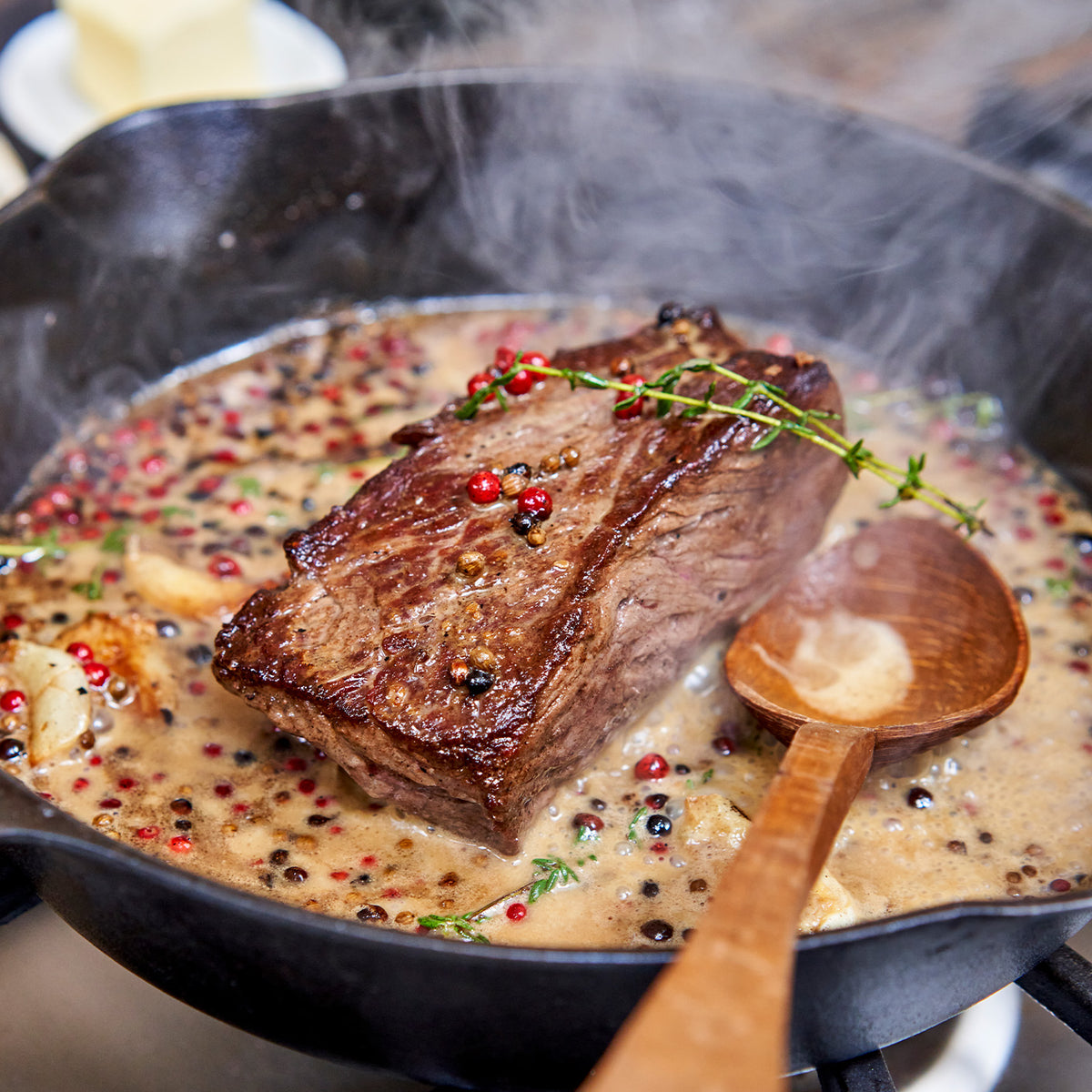 Steak being cooked in a skillet with herbs and spices, wooden spoon visible