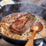Steak being cooked in a skillet with herbs and spices, wooden spoon visible