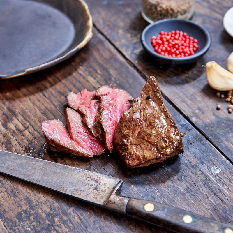 Sliced bavette steak on a wooden cutting board with a knife, surrounded by spices and seasonings.
