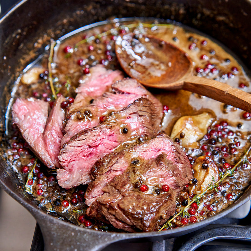 Sliced steak cooking in a pan with herbs and spices, wooden spoon visible