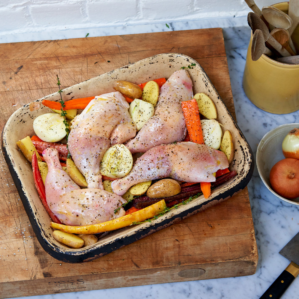 Raw chicken legs on cooking sheet surrounded by onions, potatoes, and carrots. 