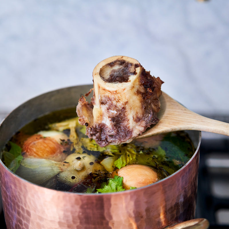 Copper pot with bone broth and a wooden spoon lifting a bone.