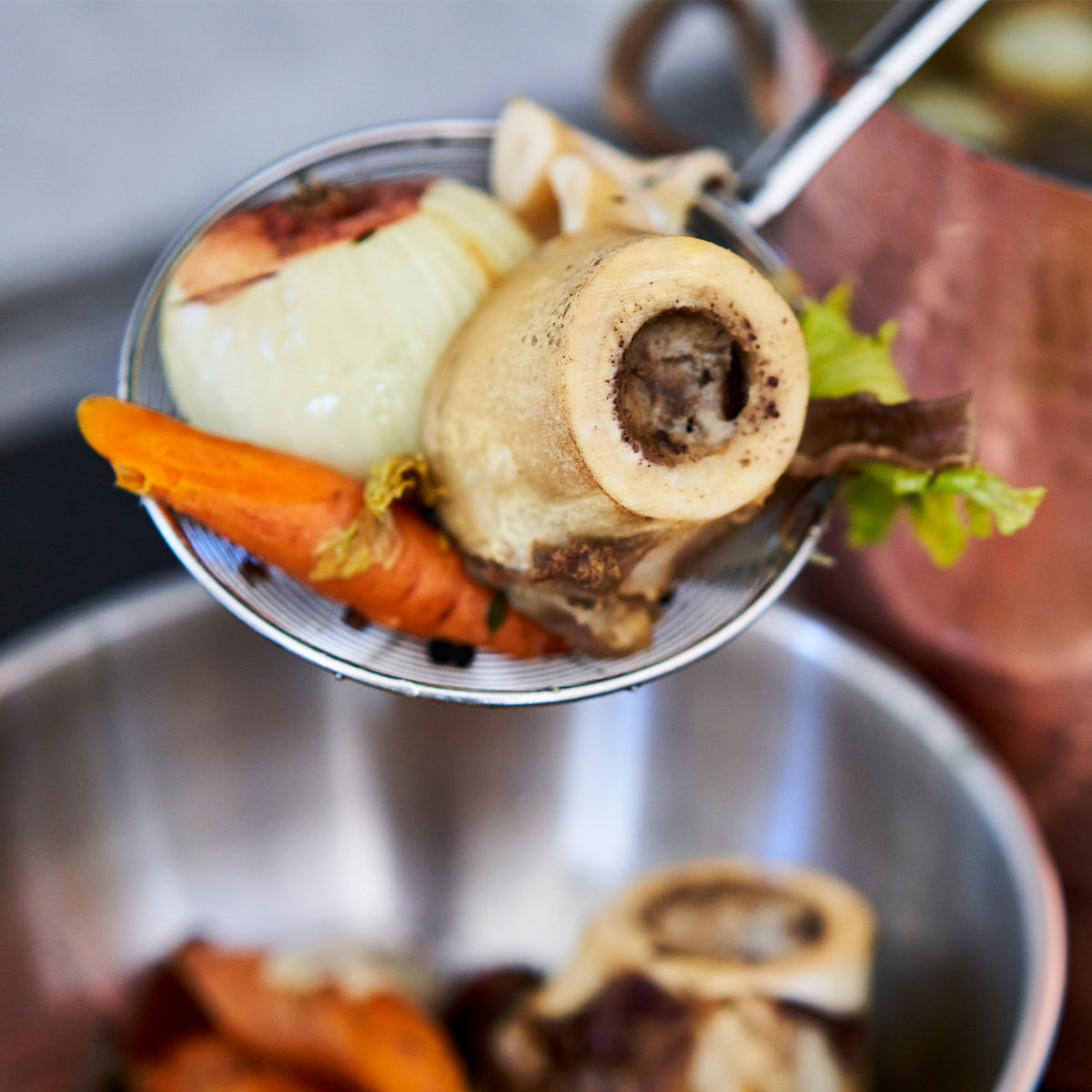 Close-up of a ladle with bones, carrots, and other ingredients in a pot.