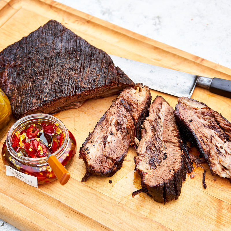 Sliced smoked brisket on a wooden cutting board with a jar of red pepper flakes and a knife.
