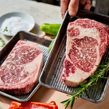 Two raw steaks on metal trays with herbs and vegetables on a wooden surface.