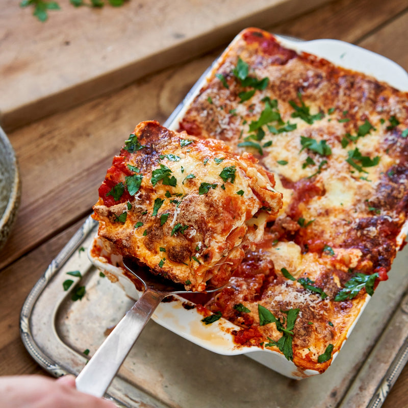 Baked lasagna with a serving being lifted, garnished with parsley on a wooden surface.