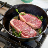 Two raw steaks with rosemary in a cast iron skillet on a stovetop.