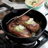 Two steaks with green compound butter being cooked in a black skillet on a stovetop.