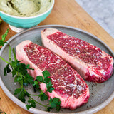 Two raw steaks on a plate with a bowl of green herb mixture on a wooden surface.