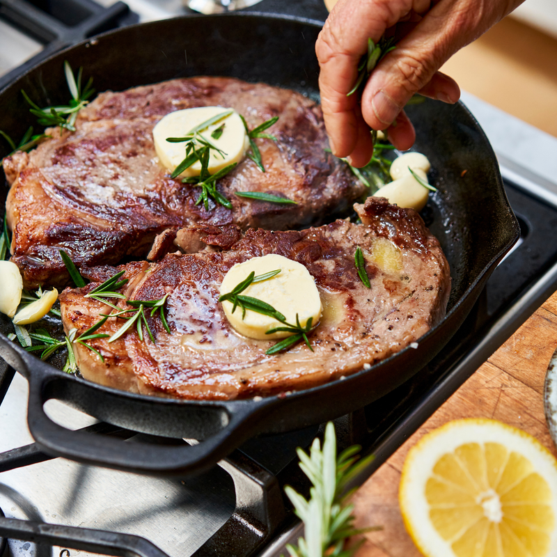 Two seared steaks in a cast iron skillet with butter and herbs, on a stove top.