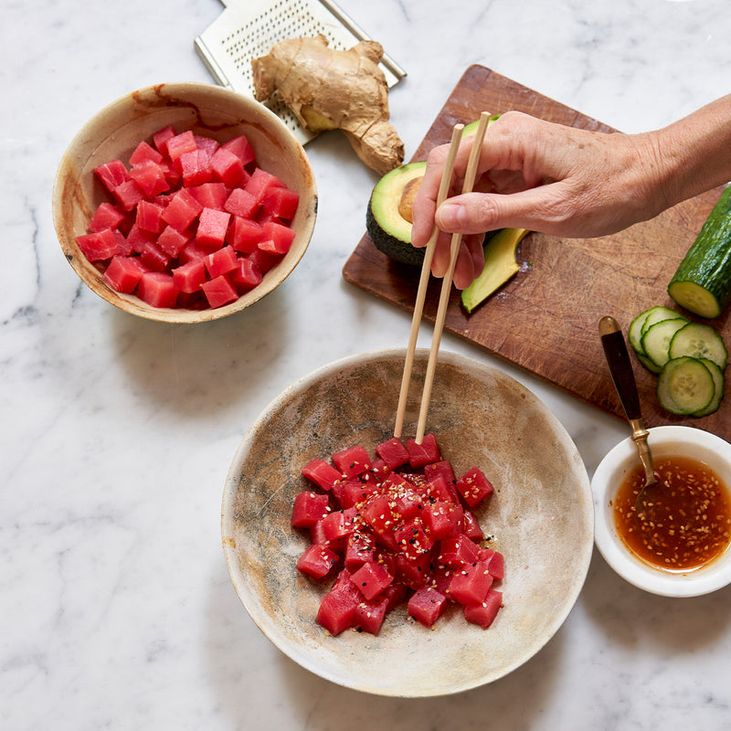 Tuna Poke Bowl being enjoyed with chopsticks. Avocado, cucumber, ginger and avocado are on cutting board.