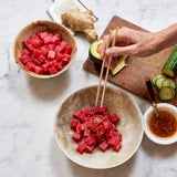 Tuna Poke Bowl being enjoyed with chopsticks. Avocado, cucumber, ginger and avocado are on cutting board.